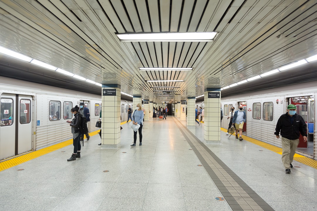 On the platform for Bloor-Yonge Station. Subway trains have arrived on each side of the platform. The left side train is still closed and passengers are waiting to board. The right side train is open and passengers are exiting the train.